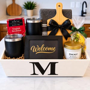 May include: A white wooden gift crate with a black letter M, containing two black tumblers, English Breakfast tea, a dark gray waffle towel, a bamboo cutting board with a black bow, a black Welcome sign, and a DKNY candle with a bag of lemons.
