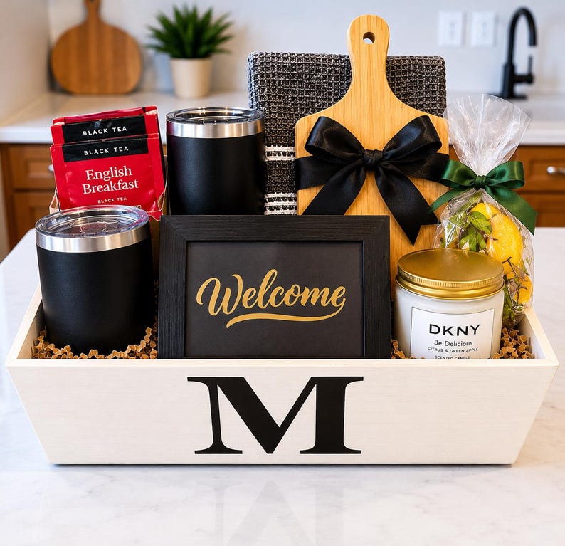 May include: A white wooden gift crate with a black letter M, containing two black tumblers, English Breakfast tea, a dark gray waffle towel, a bamboo cutting board with a black bow, a black Welcome sign, and a DKNY candle with a bag of lemons.