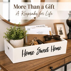 May include: A rectangular white wooden storage tray with cut-out handles and the black text Home Sweet Home printed on the front. The tray contains a small potted green plant, envelopes, a set of keys with a wooden tag, and black sunglasses.