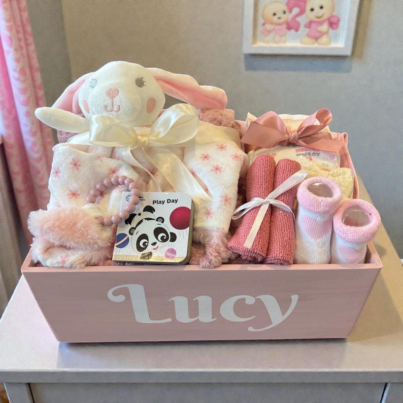 Peut inclure: Un panier cadeau en bois rose clair avec le pr&eacute;nom Lucy imprim&eacute; en blanc sur le devant. Le panier contient un lapin en peluche blanc avec un ruban cr&egrave;me, une couverture rose &agrave; motifs, un livre pour b&eacute;b&eacute; panda, un anneau de dentition et des chaussettes.
