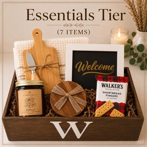 May include: A dark wooden gift crate with a white letter W containing a vanilla candle, a small bamboo cutting board with a spreader, a round wooden box, a white framed sign with the word Welcome, and a package of Walker's shortbread fingers.