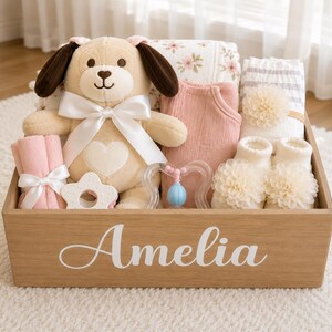 Peut inclure: Une caisse cadeau en bois avec le pr&eacute;nom Amelia imprim&eacute; en blanc sur le devant. &Agrave; l'int&eacute;rieur se trouvent un chien en peluche beige avec un n&oelig;ud blanc, un body rose, une couverture &agrave; fleurs, un anneau de dentition &eacute;toile et des chaussons.