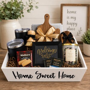 May include: A white wooden gift crate labeled Home Sweet Home containing two black tumblers, a wooden cutting board with a gold ribbon, a scrub brush, a black framed sign reading Welcome, a candle, Walkers shortbread, and Bigelow tea bags.