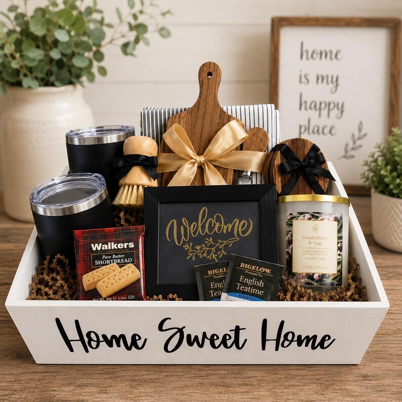 May include: A white wooden gift crate labeled Home Sweet Home containing two black tumblers, a wooden cutting board with a gold ribbon, a scrub brush, a black framed sign reading Welcome, a candle, Walkers shortbread, and Bigelow tea bags.