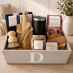 May include: A gray wooden gift crate with a white letter D containing a navy and white striped towel, a wooden cutting board, a wooden scrub brush, a black tumbler, a candle labeled Coconut Oak & Amber, Harry & David truffles, and soap.