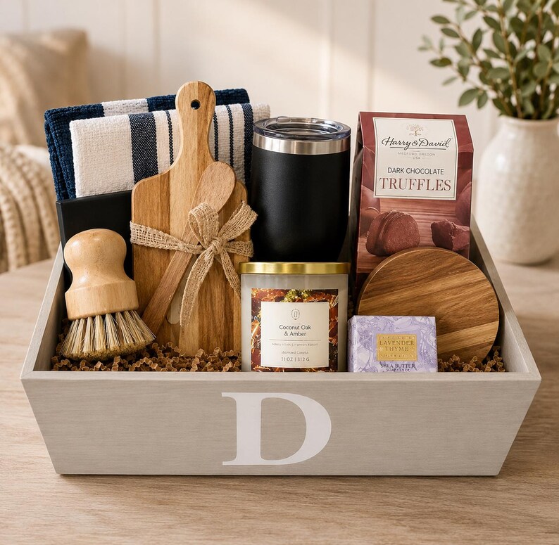 May include: A gray wooden gift crate with a white letter D containing a navy and white striped towel, a wooden cutting board, a wooden scrub brush, a black tumbler, a candle labeled Coconut Oak & Amber, Harry & David truffles, and soap.