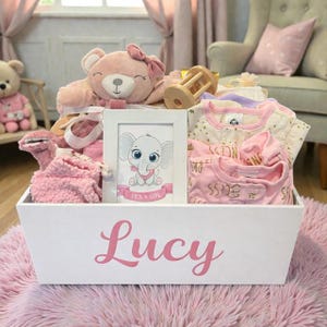 Peut inclure: Un panier cadeau en bois blanc avec le pr&eacute;nom Lucy imprim&eacute; en &eacute;criture rose. Le panier contient un ours en peluche rose, un hochet en bois, un cadre photo blanc avec une illustration d'&eacute;l&eacute;phant et le texte It's a Girl, et des v&ecirc;tements pour b&eacute;b&eacute;.