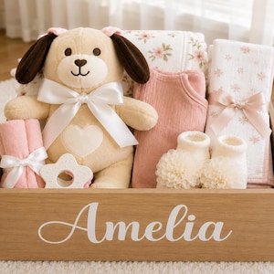 May include: A wooden gift box with the name Amelia printed in white script on the front. Inside are a tan plush dog with a white bow, a pink onesie, a white star-shaped teether, a pair of cream booties, and two folded blankets with floral prints.