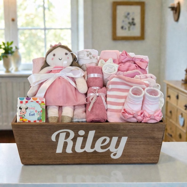 Peut inclure: Un panier cadeau en bois avec le nom Riley imprim&eacute; en blanc, contenant une poup&eacute;e en peluche en robe rose, un petit livre intitul&eacute; My Baby Loves to Eat, une serviette rose, des v&ecirc;tements ray&eacute;s rose et blanc et des chaussons roses avec des n&oelig;uds.
