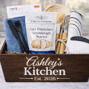 May include: A wooden gift basket filled with kitchen items. Includes a sourdough starter, utensils, a blue cloth, and a white oven mitt. The basket is labeled "Ashley's Kitchen Est. 2026".