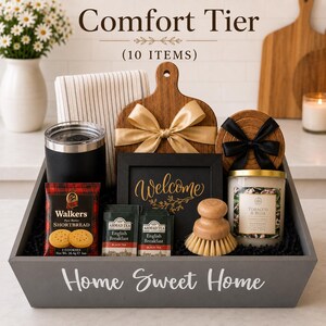 May include: A gray wooden gift crate labeled Home Sweet Home containing ten items: a black tumbler, a striped towel, a wooden cutting board with a gold bow, a Welcome sign, two Ahmad Tea bags, Walkers shortbread, a wooden brush, and a Tobacco & Rose candle.