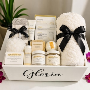 May include: A white wooden gift crate labeled Gloria containing a white plush towel with a black ribbon, a gray and white striped towel, a white loofah, a scented candle, fragrance oil, salt scrub, vanilla body butter, soap, and Sadoer face masks.