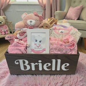 Peut inclure: Un panier cadeau en bois fonc&eacute; avec le nom Brielle en lettres blanches, contenant un ours en peluche rose avec un n&oelig;ud, un hochet en bois, un cadre blanc avec une image d'&eacute;l&eacute;phant et le texte It's a Girl, et des v&ecirc;tements de b&eacute;b&eacute; pli&eacute;s.