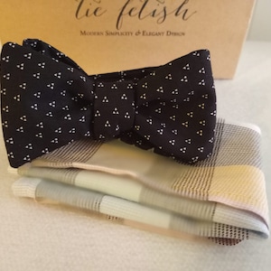Könnte beinhalten: Eine schwarze Fliege mit einem weißen Dreiecksmuster und ein passendes Einstecktuch mit einem hellblauen, beige und braunen Karomuster.