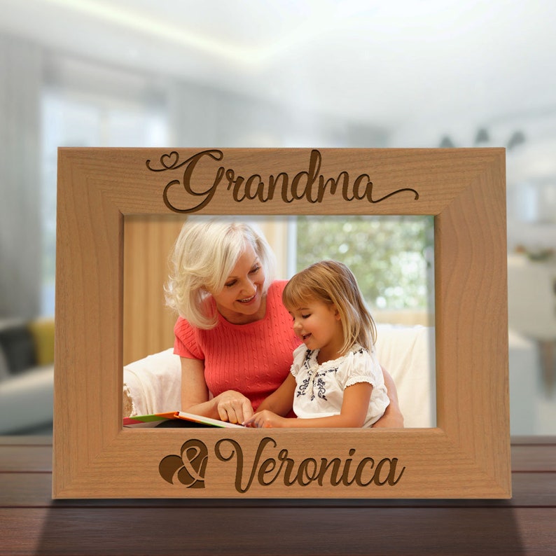 May include: A wooden picture frame with a light brown finish. The frame has a cutout design with the words "Grandma" and "Veronica" and two hearts. The frame is holding a photo of an older woman and a young girl.