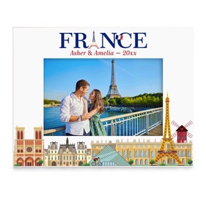 May include: A white photo frame with a blue and red "FRANCE" title and a picture of a couple in front of the Eiffel Tower. The frame also features a cityscape of Paris with iconic landmarks like the Eiffel Tower, Notre Dame Cathedral, and the Louvre Museum.