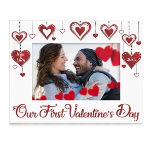 May include: White photo frame with a red heart design. The frame has a photo of a couple smiling at each other. The text "Our First Valentine's Day" is written in red cursive at the bottom of the frame. The text "Ivan & Lucy" is written in white on a red heart at the top left of the frame. The text "20xx" is written in white on a red heart at the top right of the frame.