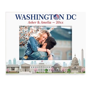 May include: A white photo frame with a picture of a couple embracing in front of a cherry blossom tree. The frame has a blue and red design with the text "WASHINGTON DC" and "Asher & Amelia ~ 20xx" at the top. The bottom of the frame features a cityscape of Washington D.C. with iconic landmarks like the White House, the Capitol Building, and the Washington Monument.