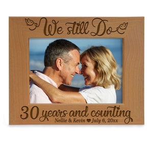 May include: A wooden photo frame with a light brown finish. The frame has a cutout design that says "We still do" with two birds on either side. The bottom of the frame says "30 years and counting" with the names "Nellie & Kevin" and the date "July 6, 20xx".