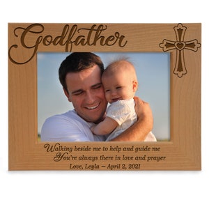 May include: A wooden picture frame with the word "Godfather" engraved on the top. The frame has a cross with a heart in the center on the right side. The frame has a photo of a man holding a baby. The text below the photo reads "Walking beside me to help and guide me You're always there in love and prayer Love, Leyla - April 2, 2021"