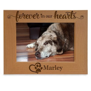 May include: Wooden photo frame with a light brown finish. The frame has a cutout design that says "forever in our hearts" with a small heart on each side. The frame also has a cutout design of a heart with a paw print inside and the name "Marley" below it. The frame has a picture of a brown and white dog inside.