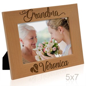 May include: A wooden picture frame with a 5x7 inch opening. The frame is engraved with the words "Grandma & Veronica" and a heart symbol. The frame is light brown in color.