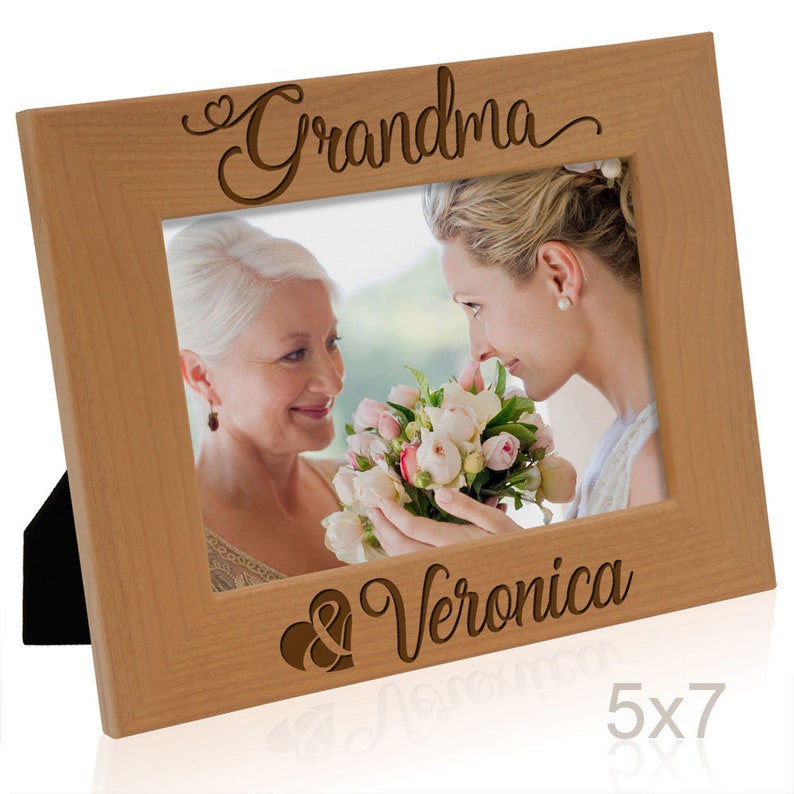 May include: A wooden picture frame with a 5x7 inch opening. The frame is engraved with the words "Grandma & Veronica" and a heart symbol. The frame is light brown in color.