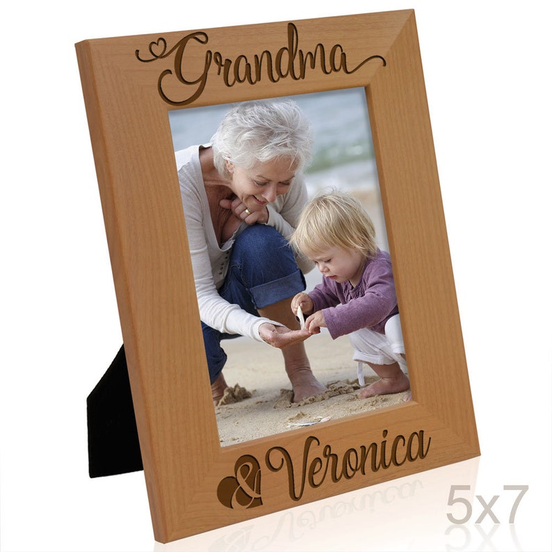 May include: A wooden picture frame with a light brown finish. The frame has a cutout design with the words "Grandma & Veronica" and a heart symbol. The frame is designed to hold a 5x7 inch photo.