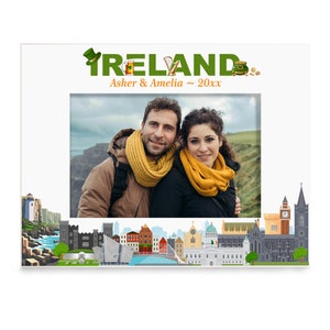 May include: A white picture frame with a green and gold "IRELAND" title and a couple's photo inside. The frame features a cityscape of Dublin, Ireland, with iconic landmarks like Trinity College and the Ha'penny Bridge.