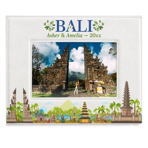 May include: White photo frame with a picture of a traditional Balinese gate. The frame has a white background with the words "BALI" in blue letters and "Asher & Amelia ~ 20xx" in black letters. The frame is decorated with a green and white floral design.