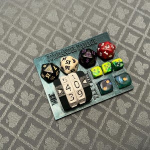 May include: A teal rectangular tracker board with the text COMMANDER DAMAGE and LIFE TAX printed on it. The board features a black three-digit mechanical counter displaying 5409 and includes various multi-sided dice in red, black, white, and green.