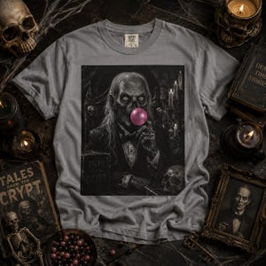 May include: A grey short-sleeve t-shirt featuring a graphic print of a skeletal figure in a suit blowing a pink bubble. The figure sits in a dark room with candles and a book titled Tales from the Crypt. The shirt has a Comfort Colors brand label.
