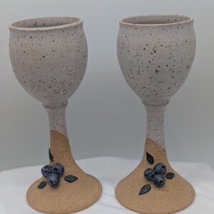 May include: Two ceramic goblets featuring a speckled off-white bowl and a tan base. The stem of each goblet is decorated with a cluster of three-dimensional dark blue blueberries and small green leaves attached to the lower section of the stem.