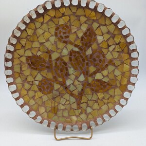 May include: A circular mosaic plate featuring a floral pattern in shades of yellow and brown. The plate has a terracotta-colored base with a border of white glass tiles. The center displays a stylized flower design made of small, irregular glass pieces.