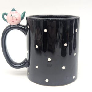 May include: Black ceramic mug with white polka dots. The handle features a decorative pink teapot with green accents and cherries. The mug is cylindrical and suitable for hot or cold beverages.
