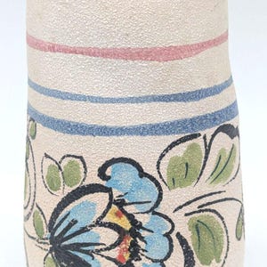 May include: A cream ceramic vase with a textured finish and a scalloped rim. It features blue and pink horizontal stripes and a floral design with blue flowers and green leaves. The vase is approximately 15 cm tall.