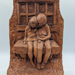 May include: A brown sculpture depicting two figures seated on a bench, with a window in the background. The figures are close together, suggesting intimacy. The sculpture has a textured surface and a rustic appearance.