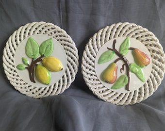 May include: Two round, white woven wall plaques. Each plaque features a central, circular ceramic design with a fruit motif. One has yellow lemons, the other has orange pears, both with green leaves and brown branches.