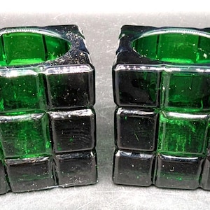 May include: Two square candle holders with a dark green glass design. Each holder is made of small, square glass blocks, creating a textured, geometric pattern. The top of each holder has a circular opening for a candle.
