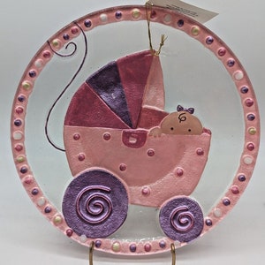 May include: A round, pink baby-themed decorative plate. The plate features a baby carriage with a baby inside, all in shades of pink and purple. The plate's border is decorated with pink and white dots. A gold string is attached to the top.