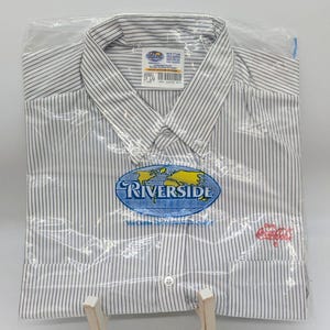 May include: A new, white button-down shirt with thin, vertical black stripes, packaged in clear plastic. The shirt features a "Riverside" logo with a world map and the Coca-Cola logo embroidered in red.