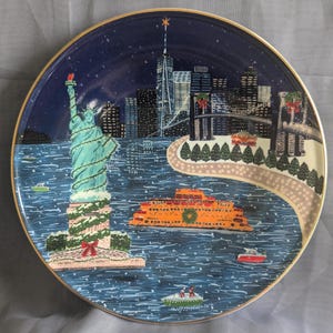 May include: Decorative plate featuring a winter scene of New York City. The Statue of Liberty is adorned with garland. An orange ferry boat sails on the water. The plate has a gold rim and a dark blue background with a starry night sky.