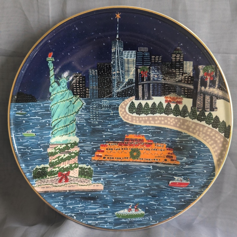 May include: Decorative plate featuring a winter scene of New York City. The Statue of Liberty is adorned with garland. An orange ferry boat sails on the water. The plate has a gold rim and a dark blue background with a starry night sky.