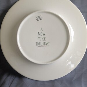 May include: White ceramic plate with the text "A NEW YORK HOLIDAY" in black lettering. The plate is from the brand "ANTHROPOLOGIE". The plate is round with a slightly raised edge.