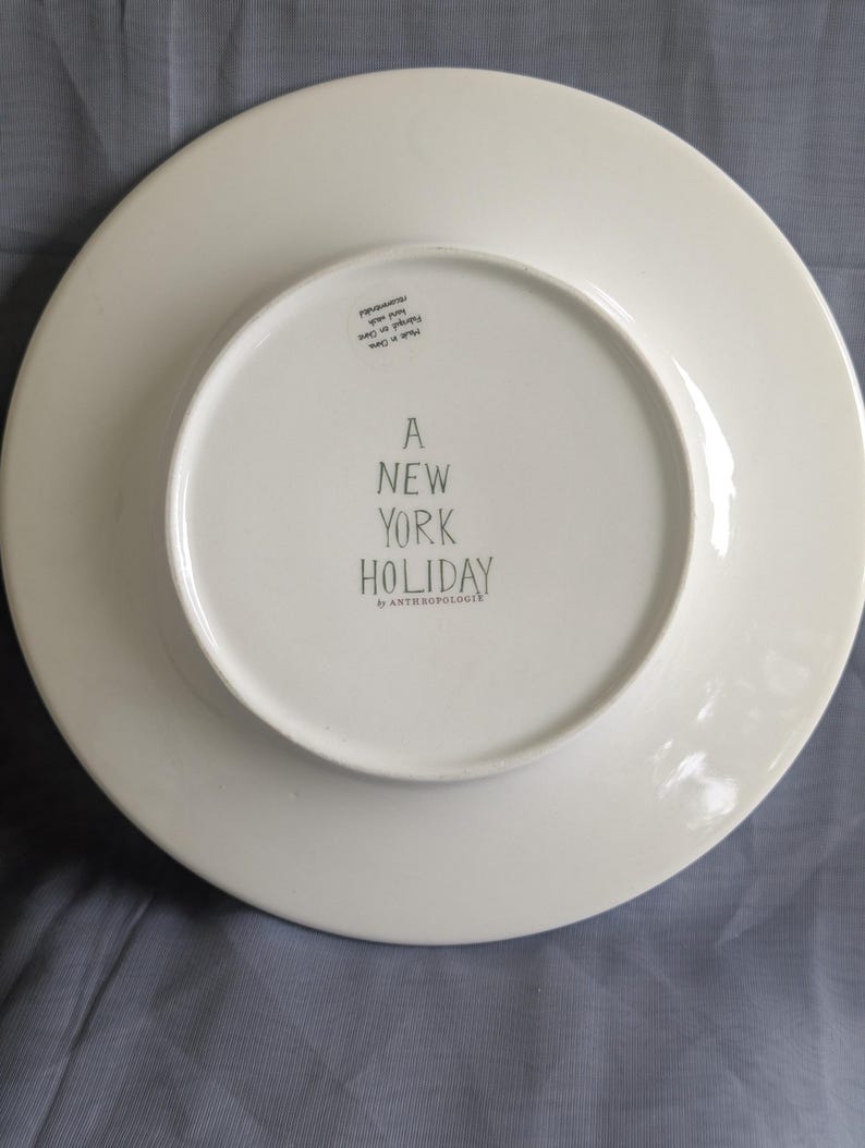 May include: White ceramic plate with the text "A NEW YORK HOLIDAY" in black lettering. The plate is from the brand "ANTHROPOLOGIE". The plate is round with a slightly raised edge.