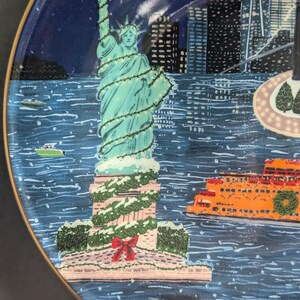 May include: A decorative plate featuring a winter scene of the Statue of Liberty in New York City. The statue is decorated with lights and snow, with an orange ferry and city skyline in the background. The plate has a gold rim.