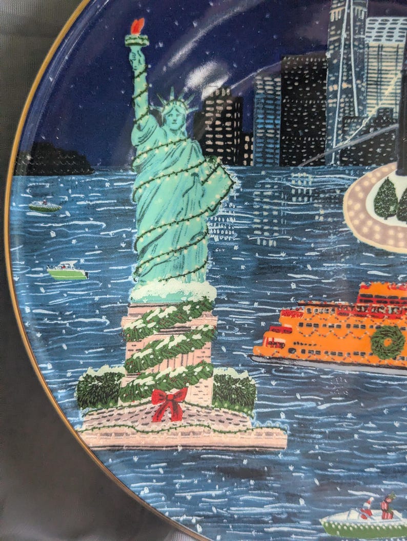 May include: A decorative plate featuring a winter scene of the Statue of Liberty in New York City. The statue is decorated with lights and snow, with an orange ferry and city skyline in the background. The plate has a gold rim.