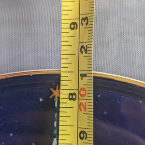 May include: A yellow measuring tape shows a plate's diameter. The plate has a dark blue background with a gold rim and a design featuring a building and stars. The tape measures approximately 8 inches.