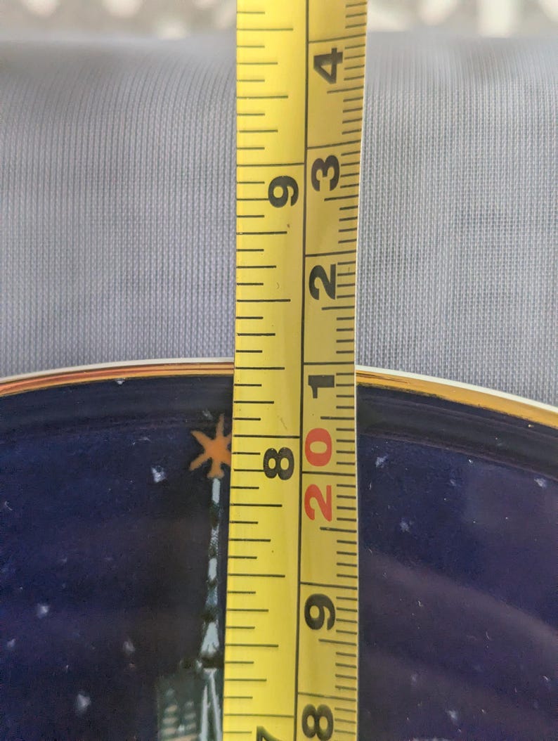 May include: A yellow measuring tape shows a plate's diameter. The plate has a dark blue background with a gold rim and a design featuring a building and stars. The tape measures approximately 8 inches.