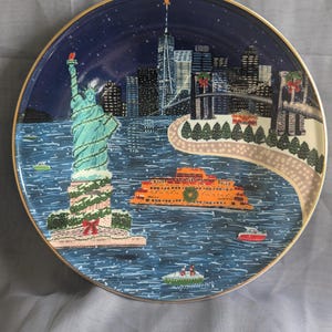 May include: A decorative plate featuring a winter scene of New York City. The plate depicts the Statue of Liberty, the city skyline, and an orange ferry boat on a blue background. The plate has a gold rim.